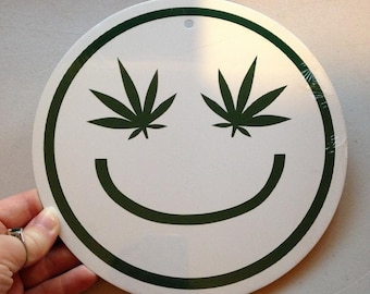 Op de afbeelding: Een rond wit bord met een groene rand en een smiley. De ogen van de smiley zijn groene marihuanabladeren.