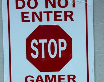 May include: White rectangular sign with a red border. The sign reads "DO NOT ENTER" above a red octagon with the word "STOP" in white. Below the octagon, the sign says "GAMER AT PLAY".