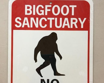 Op de afbeelding: Een wit metalen bord met een rode rand en een zwart silhouet van Bigfoot die loopt. Het bord staat er "Bigfoot Sanctuary No Hunting" op.