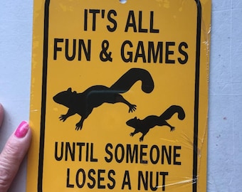 May include: Yellow metal sign with black text and images of squirrels. The sign reads "It's all fun & games until someone loses a nut."