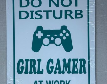 May include: A rectangular white metal sign with rounded corners, featuring the text "DO NOT DISTURB" above a teal controller graphic and the words "GIRL GAMER AT WORK". The sign has a hole at the top for hanging.
