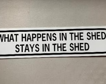 May include: A rectangular white sign with black text that reads "WHAT HAPPENS IN THE SHED STAYS IN THE SHED." The sign has a black border and is likely made of metal or a similar material.