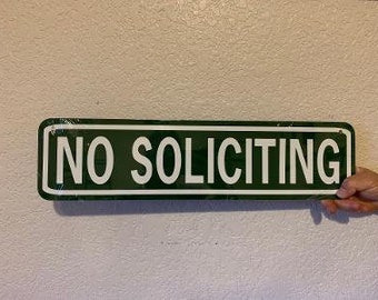 Op de afbeelding: Een groen metalen bord met witte letters met de tekst "NO SOLICITING".