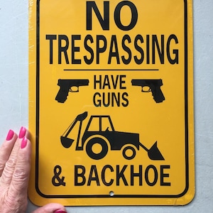 May include: Yellow metal sign with black text that reads "NO TRESPASSING HAVE GUNS & BACKHOE". The sign also features two black silhouettes of handguns and a black silhouette of a backhoe.