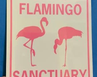May include: White metal sign with pink text that reads "LAWN FLAMINGO SANCTUARY". The sign features two pink flamingo silhouettes.