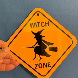 May include: A yellow diamond-shaped sign with black text and a silhouette of a witch flying on a broomstick. The sign reads "Witch Zone".