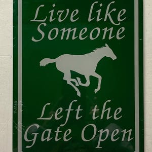 以下が含まれることがあります： 「Live like someone left the gate open」という白い文字が書かれた緑色の金属製の看板。看板の中央には、走っている馬の白いシルエットがあります。