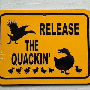 Può includere: Cartello rettangolare giallo con angoli arrotondati e bordo nero. Il cartello presenta una silhouette nera di un'anatra in volo, le parole "RELEASE THE QUACKIN'" e sagome di un'anatra e anatroccoli.