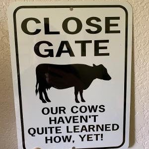 Op de afbeelding: Een wit metalen bord met zwarte tekst die "CLOSE GATE OUR COWS HAVEN'T QUITE LEARNED HOW, YET!" luidt en een zwart silhouet van een koe.