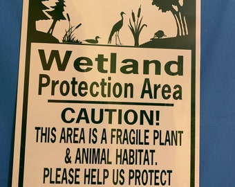 May include: A white sign with green trim and a black hole in the top left corner. The sign has a green background with a white silhouette of a wetland scene. The text on the sign reads "Wetland Protection Area CAUTION! This area is a fragile plant & animal habitat. Please help us protect this natural resource Do Not Disturb."