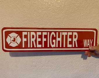 May include: Red metal sign with white text that reads "FIREFIGHTER WAY". A white firefighter symbol is on the left side of the sign.