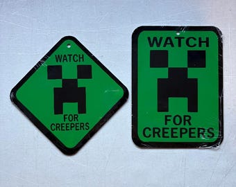 May include: Two green signs with black text and a pixelated face design. The signs read "WATCH FOR CREEPERS". One sign is diamond-shaped, the other is rectangular. Both have a hole at the top for hanging.
