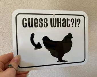 May include: White metal sign with black text and a silhouette of a chicken. The text reads "Guess What?!" with an arrow pointing to the chicken's rear end.