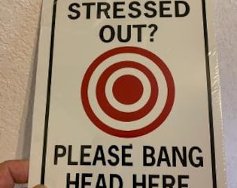 May include: White sign with black border and text that reads "Stressed Out?" with a red and white target graphic and the text "Please Bang Head Here".