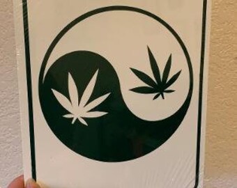 Op de afbeelding: Een rechthoekig wit bord met een zwarte rand heeft een yin-yang ontwerp. Eén kant is donkergroen met een cannabisblad, de andere wit met een cannabisblad. Het bord wordt door een hand vastgehouden.