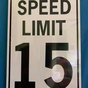 Op de afbeelding: Een wit metalen bord met zwarte tekst en cijfers. Het bord geeft "SPEED LIMIT 15" aan.