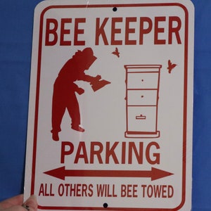 May include: A white metal sign with red text and a red silhouette of a beekeeper. The sign reads "Bee Keeper Parking" with an arrow pointing to the left and an arrow pointing to the right. The text below the arrows reads "All others will bee towed".