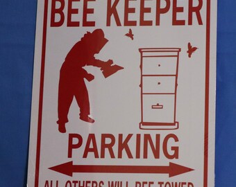 Op de afbeelding: Een wit metalen bord met rode tekst en een rood silhouet van een imker. Het bord staat "Bee Keeper Parking" met een pijl naar links en een pijl naar rechts. De tekst onder de pijlen staat "All others will bee towed".