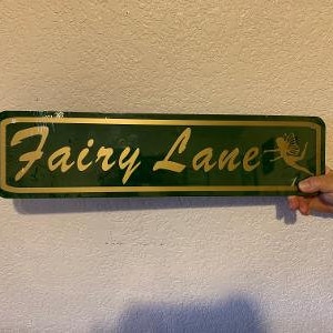 Puede incluir: Un letrero de metal verde con letras doradas que dice "Fairy Lane". Una silueta de hada dorada está en el lado derecho del letrero.