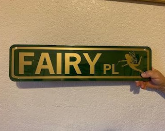 Op de afbeelding: Een groen metalen bord met gouden letters met de tekst "FAIRY PL" en een gouden fee-silhouet aan de rechterkant.