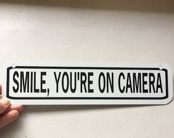 Op de afbeelding: Een wit rechthoekig bord met een zwarte rand en zwarte tekst die "SMILE, YOU'RE ON CAMERA" luidt.