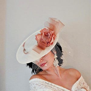 Peut inclure: Un chapeau blanc avec une grande fleur rose et un voile. Le chapeau est d&eacute;cor&eacute; d'un ruban brun.