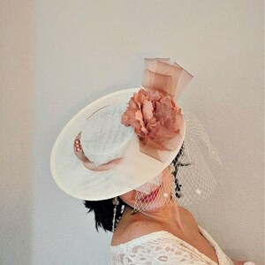 Peut inclure: Un chapeau blanc avec une grande fleur rose et un voile. Le chapeau est en paille et a une large bordure. La fleur est en tissu et a une texture pliss&eacute;e. Le voile est en tulle et est attach&eacute; &agrave; l'arri&egrave;re du chapeau.