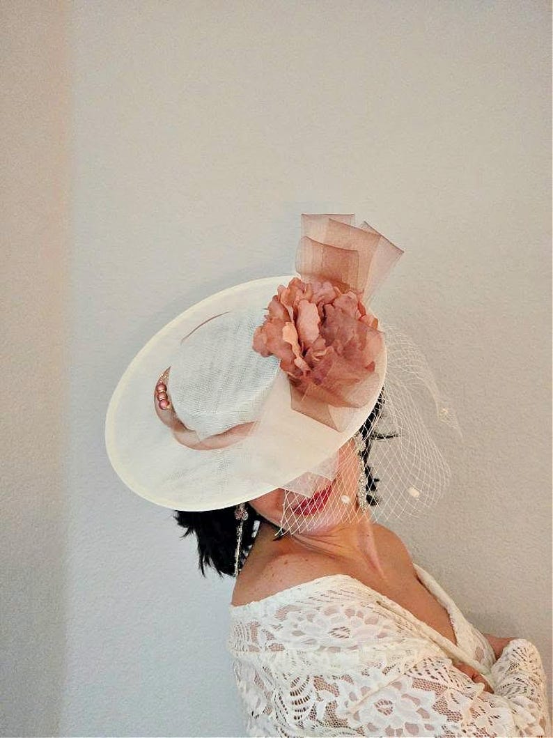 Peut inclure: Un chapeau blanc avec une grande fleur rose et un voile. Le chapeau est en paille et a une large bordure. La fleur est en tissu et a une texture pliss&eacute;e. Le voile est en tulle et est attach&eacute; &agrave; l'arri&egrave;re du chapeau.