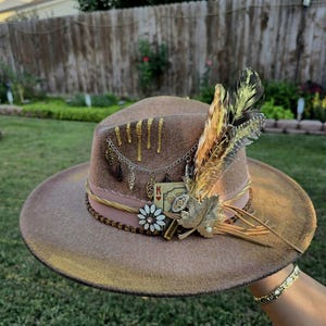 May include: A pink felt fedora hat with gold accents, a playing card, feathers, and a beaded trim. The hat has a wide brim and a tall crown.