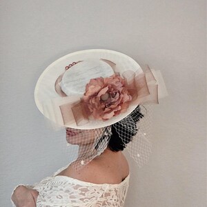 Peut inclure: Un chapeau blanc &agrave; large bord avec une fleur rose en embellissement. Le chapeau est d&eacute;cor&eacute; d'un voile blanc et d'accents de perles.