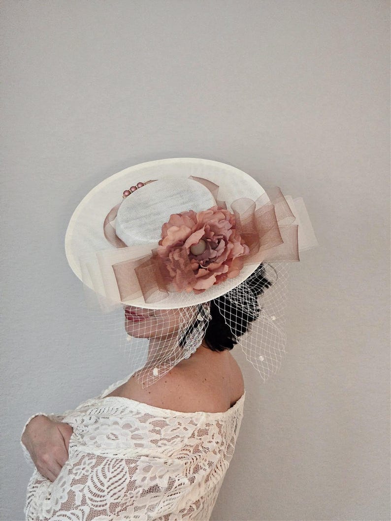 Peut inclure: Un chapeau blanc &agrave; large bord avec une fleur rose en embellissement. Le chapeau est d&eacute;cor&eacute; d'un voile blanc et d'accents de perles.