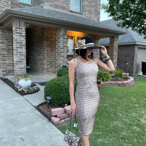 May include: A woman wearing a beige snakeskin-patterned dress, a black hat with feathers and a matching snakeskin-patterned handbag. She is standing on a sidewalk in front of a brick house.