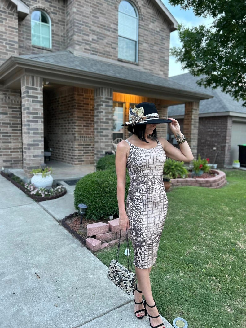 May include: A woman wearing a beige snakeskin-patterned dress, a black hat with feathers and a matching snakeskin-patterned handbag. She is standing on a sidewalk in front of a brick house.