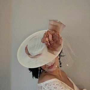 Peut inclure: Un chapeau blanc &agrave; larges bords avec une fleur brune et un voile. Le chapeau a une petite couronne et un large bord. La fleur est en tissu et a une texture pliss&eacute;e. Le voile est en filet et est attach&eacute; &agrave; l'arri&egrave;re du chapeau.
