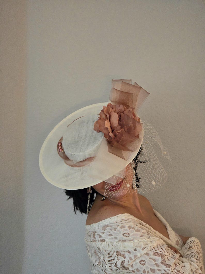 Peut inclure: Un chapeau blanc &agrave; larges bords avec une fleur brune et un voile. Le chapeau a une petite couronne et un large bord. La fleur est en tissu et a une texture pliss&eacute;e. Le voile est en filet et est attach&eacute; &agrave; l'arri&egrave;re du chapeau.