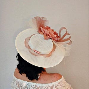 Peut inclure: Un chapeau blanc &agrave; large bord avec un accent de fleur rose. Le chapeau est d&eacute;cor&eacute; d'un ruban rose et d'un petit groupe de perles.