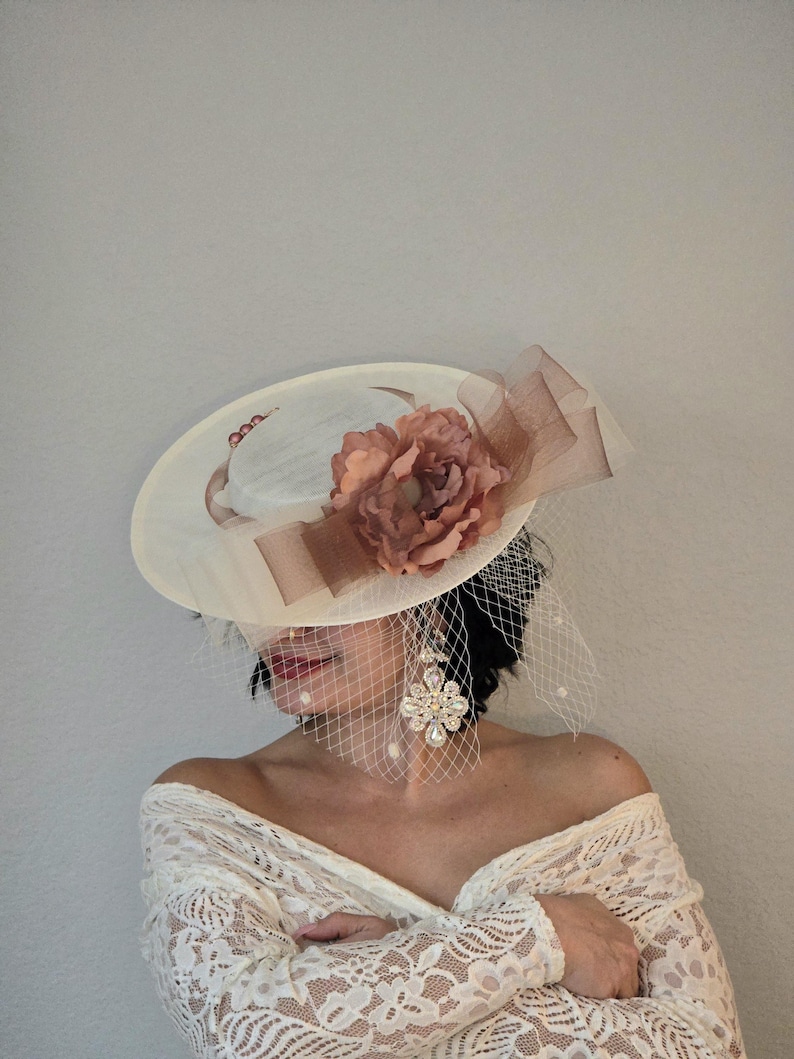 Peut inclure: Un chapeau blanc &agrave; large bord avec un accent de fleur marron. Le chapeau est d&eacute;cor&eacute; d'un voile transparent et d'un ruban marron. Le chapeau est port&eacute; par une personne aux cheveux longs et fonc&eacute;s et un haut en dentelle blanche.