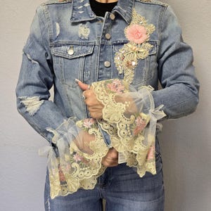 May include: Light blue denim jacket with distressed details, adorned with pink floral embellishments and intricate lace cuffs. Features a classic collar, button-up front, and a touch of vintage flair.