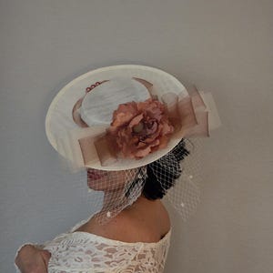 Peut inclure: Un chapeau blanc avec une grande fleur rose et un voile. Le chapeau a un large bord et un ruban en satin.