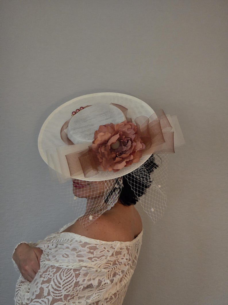 Peut inclure: Un chapeau blanc avec une grande fleur rose et un voile. Le chapeau a un large bord et un ruban en satin.
