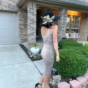 May include: A woman wearing a beige snakeskin print midi dress with a black hat and black heels. She is carrying a small beige snakeskin print handbag with a chain strap.