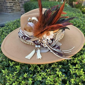 May include: A brown felt fedora hat with a wide brim, decorated with a brown and red feather, white tassels, and a patterned scarf. The hat is adorned with pearls and rhinestones.