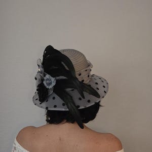 以下が含まれることがあります： 黒とグレーのドット柄の帽子に、黒い羽根と花の飾りがある。帽子は、黒髪の人の頭にかぶっている。