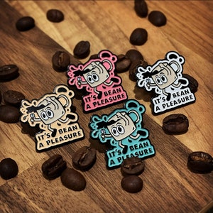 May include: Four enamel pins featuring a cartoon coffee cup character with the text "It's Bean a Pleasure". The pins are in different colours: pink, blue, teal, and beige.