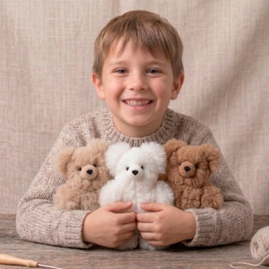 May include: A smiling child holds three plush teddy bears: one white, one light brown, and one medium brown. The child wears a light brown sweater. A ball of yarn and a small tool are on the wooden surface.