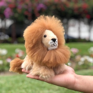 Könnte beinhalten: Ein kleiner, brauner und cremefarbener Plüschlöwe mit einer flauschigen Mähne. Der Löwe sitzt in der Hand einer Person.