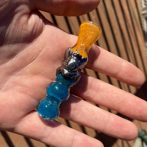 May include: A small glass pipe with a blue and orange design. The pipe has a silver metal accent in the middle.
