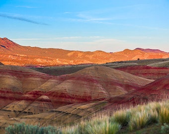 Puede incluir: Una vista panorámica de un paisaje colorido con colinas onduladas y formaciones rocosas en capas. Las colinas son una mezcla de marrón, rojo y amarillo, creando un contraste llamativo contra el cielo azul.