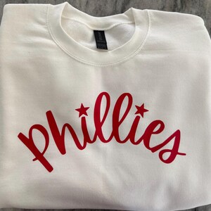 May include: A white crewneck sweatshirt with the word "Phillies" in red script across the chest. The word has two red star accents. The sweatshirt is made of a soft material and is folded neatly.