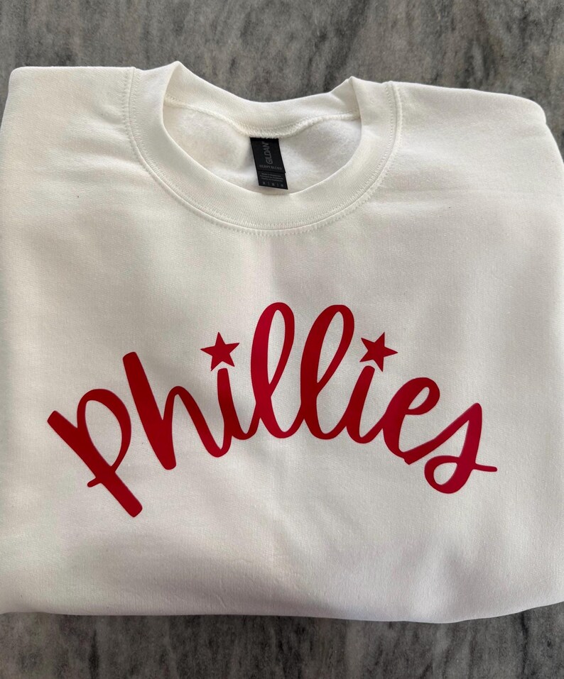 May include: A white crewneck sweatshirt with the word "Phillies" in red script across the chest. The word has two red star accents. The sweatshirt is made of a soft material and is folded neatly.